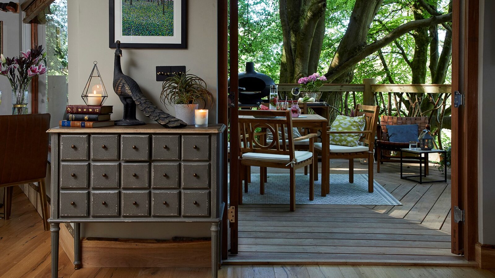 A cozy indoor-outdoor living space reminiscent of the Ravendere Treehouse, featuring a well-lit wooden deck with a dining table, chairs, and cushions under a large tree. Inside, a gray cabinet with drawers, books, and a peacock figure sits by the open glass door. Decor includes framed art and plants.