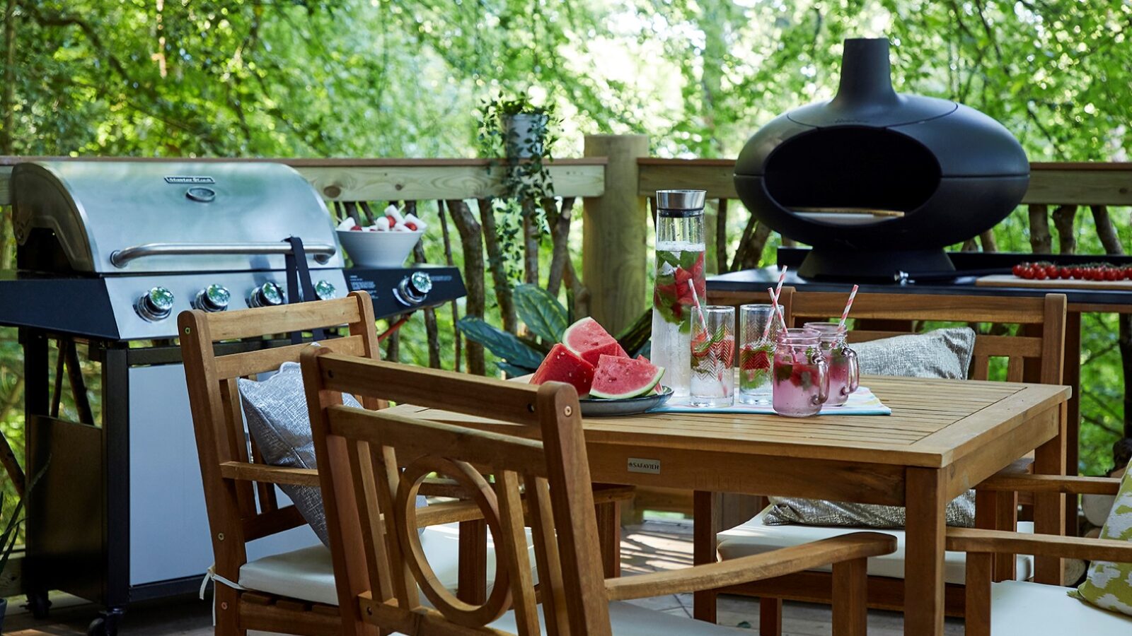 A wooden patio set for four with cushioned chairs on a deck surrounded by lush greenery evokes a Ravendere Treehouse vibe. The table holds watermelon slices, drinks, and flowers. A grill and outdoor fireplace are in the background, perfect for a summer gathering in nature.