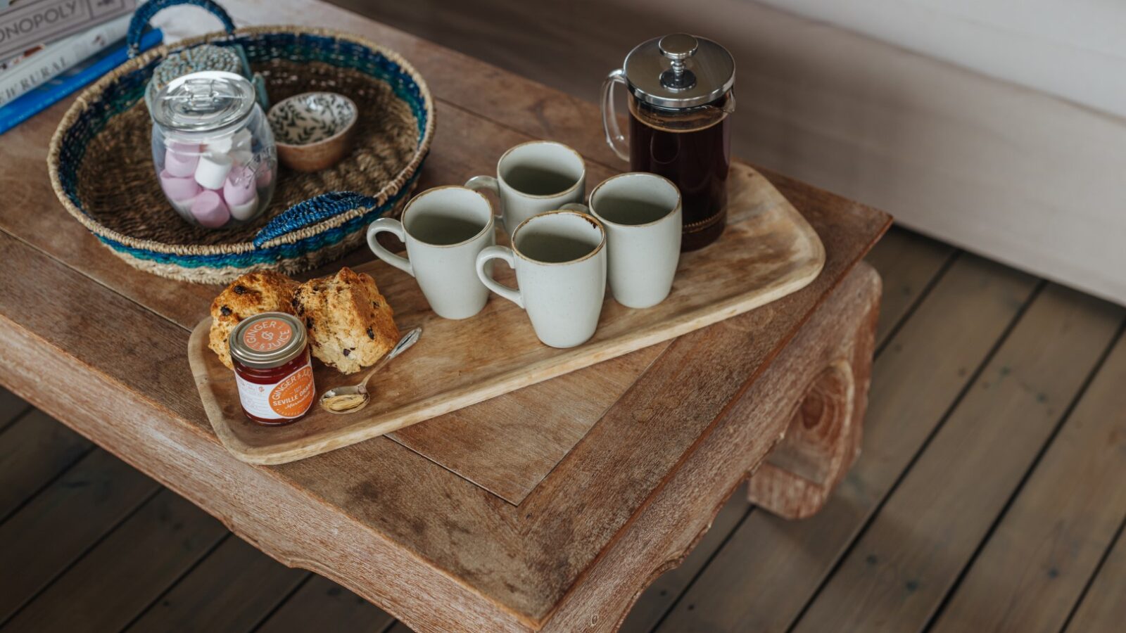 A wooden coffee table at Westgate Farm holds a tray with five white mugs and a French press filled with coffee, a small jar of marmalade, and some pastries. A woven basket with a jar of marshmallows and shells, along with board games, can also be seen on the table.