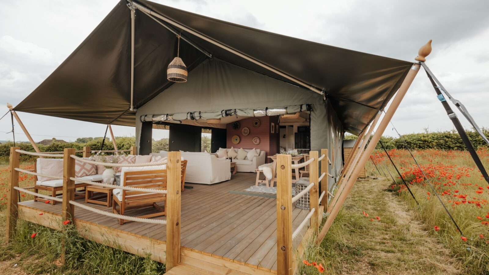 A luxurious glamping tent with modern furnishings, including white couches, a wooden table, and hanging decorations. The tent is situated on a wooden platform at Westgate Farm, surrounded by a lush field with red flowers under a cloudy sky.