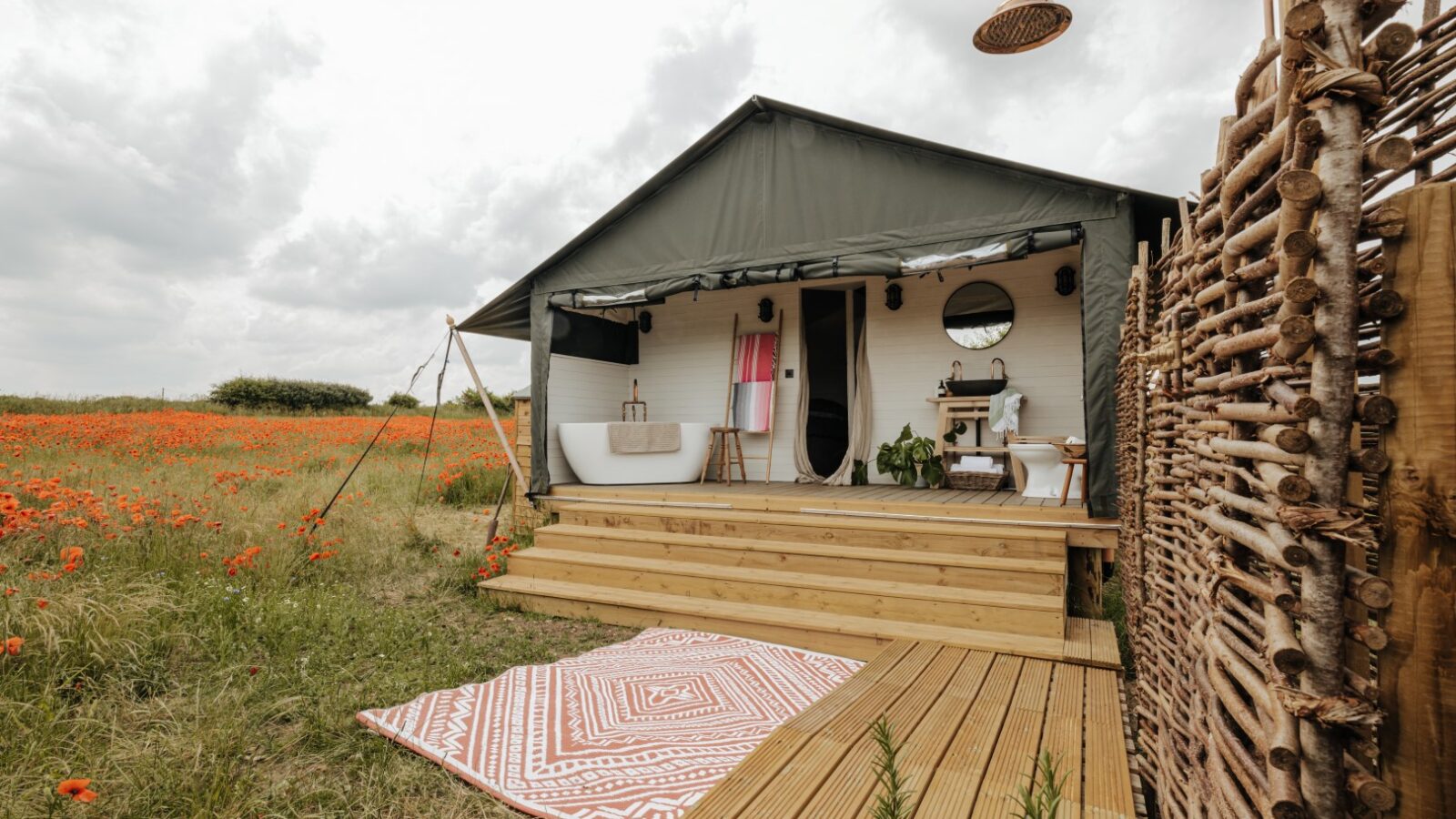 A cozy glamping tent with wooden steps and a deck is set up in a field of red poppies at Westgate Farm. The tent features a rustic design with an outdoor shower, a white bathtub, and woven privacy screens. The interior is stylishly decorated with earthy tones and a round mirror.