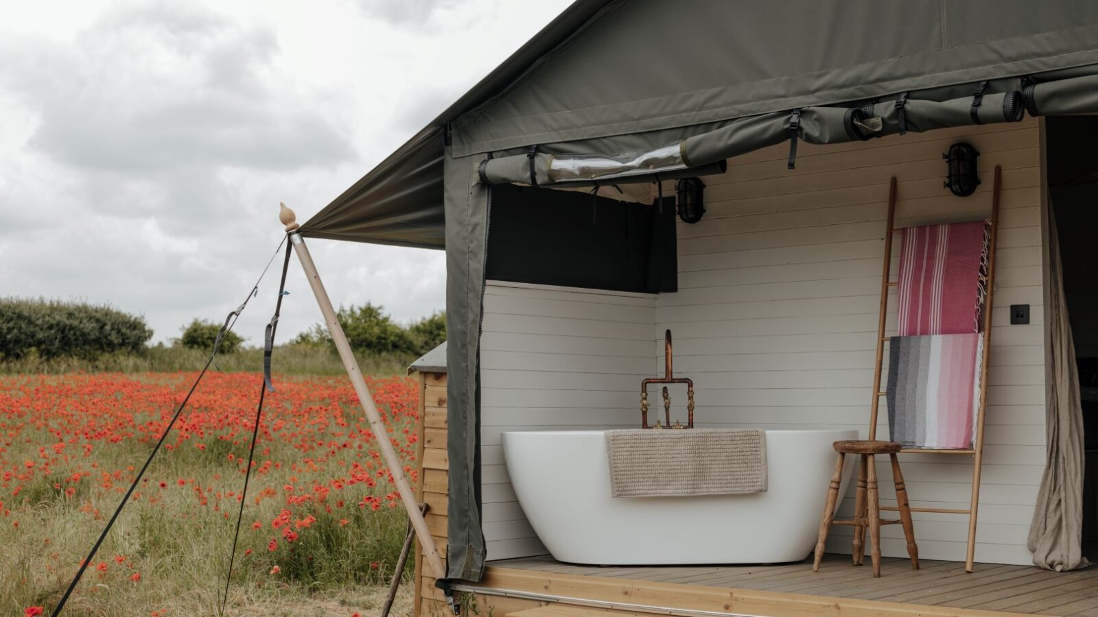 An outdoor bathtub sits on a wooden deck of a tent-like cabin at Westgate Farm, overlooking a field of red poppies. The bathtub is accompanied by a stool with a folded towel and a ladder rack holding a multicolored towel. The sky is overcast, accentuating the serene landscape.