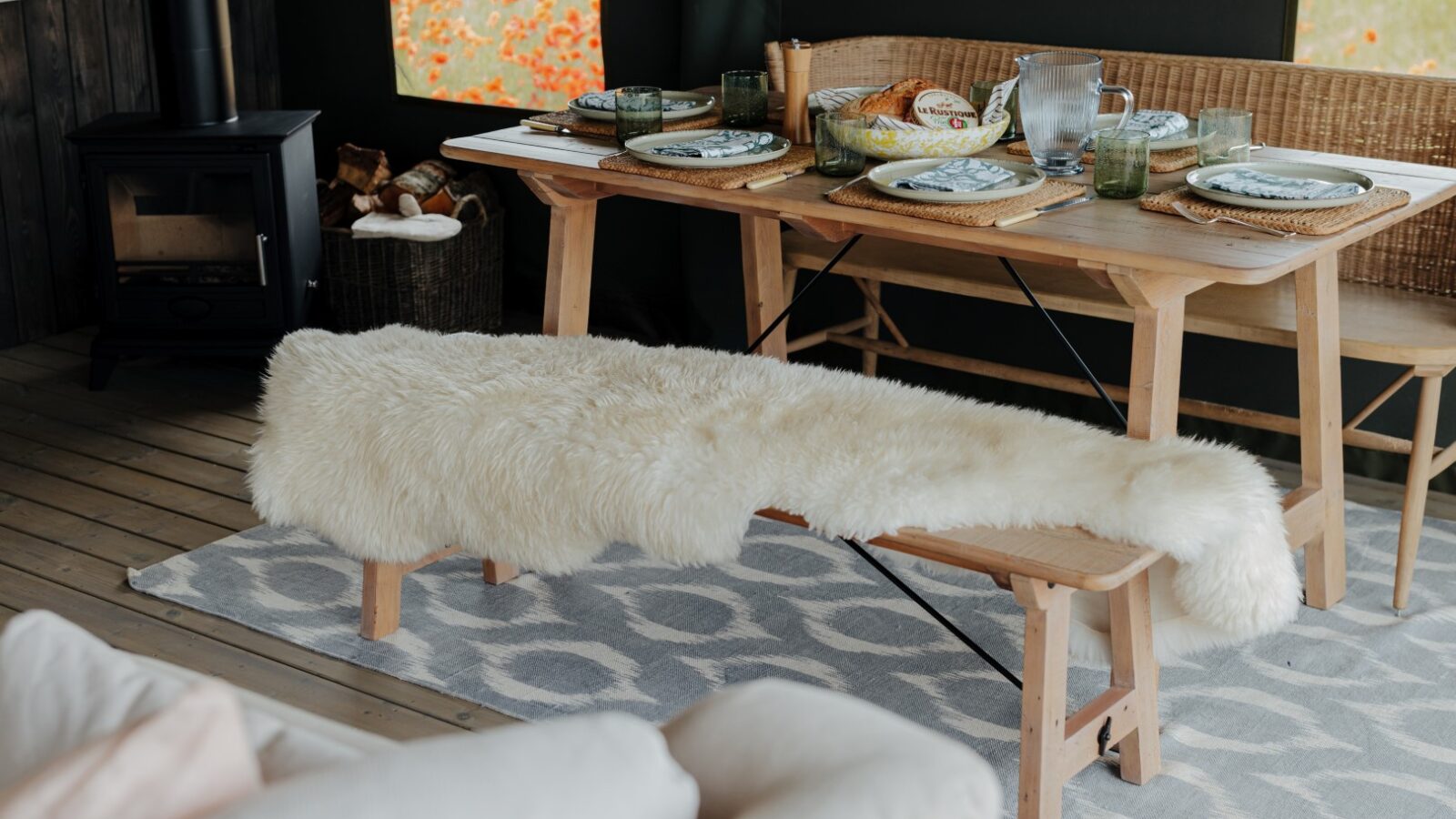 A cozy, rustic dining area at Westgate Farm features a wooden table set for a meal with plates, glasses, and a pitcher, positioned in front of a window with a view of a poppy field. A bench covered with a fluffy white fur rug sits in front of the table on a patterned rug.