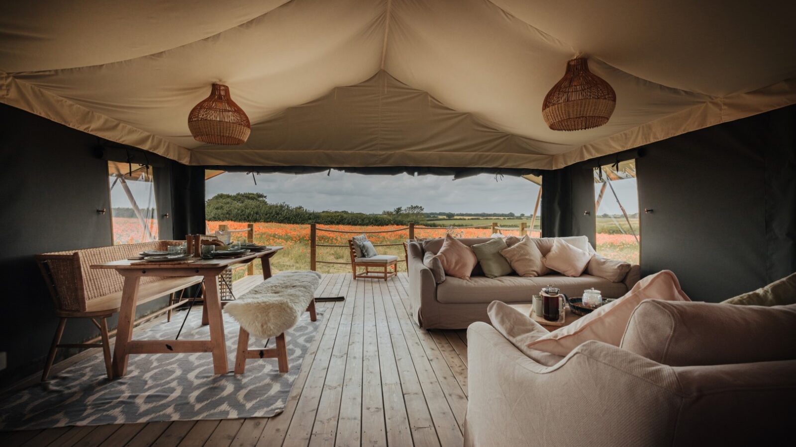 A spacious, elegantly designed glamping tent at Westgate Farm with open sides revealing a scenic view of a vibrant flower field. The interior features cozy sofas, a dining table set with dishes, wicker chairs, wooden flooring, and warm lighting, creating an inviting and comfortable ambiance.