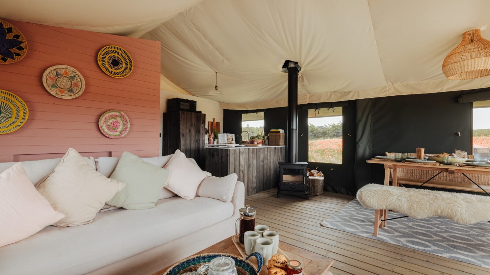 A cozy glamping tent interior at Westgate Farm features a beige sofa adorned with pillows, a wooden coffee table with snacks and drinks, a small black wood stove, and a dining area with a wooden table. Woven decor adorns the walls, while the ceiling boasts cream-colored fabric.