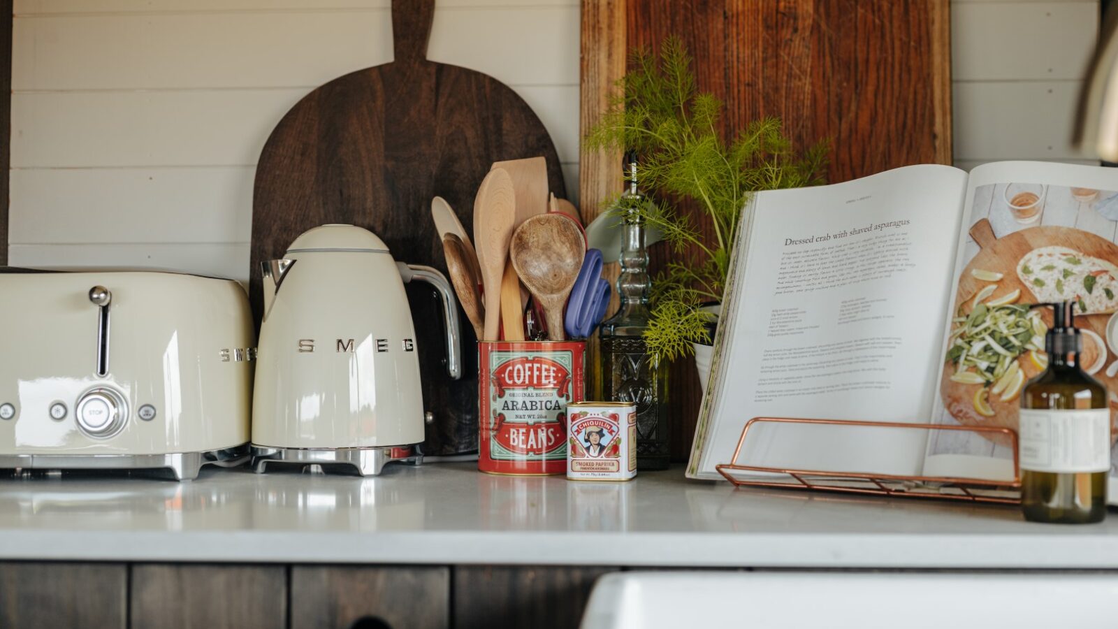 A cozy kitchen counter at Westgate Farm features a cream-colored toaster and kettle, a wooden cutting board, a container holding wooden utensils, and an open cookbook near a sprig of greenery in a glass bottle. A small vintage coffee canister adds a rustic touch.