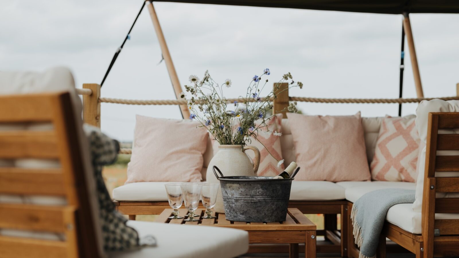 A cozy outdoor seating area at Westgate Farm features wooden furniture, cream cushions, and pink patterned pillows. A small wooden table holds a metal bucket with bottles and glasses, and a white vase with wildflowers. A green canopy provides shade overhead.