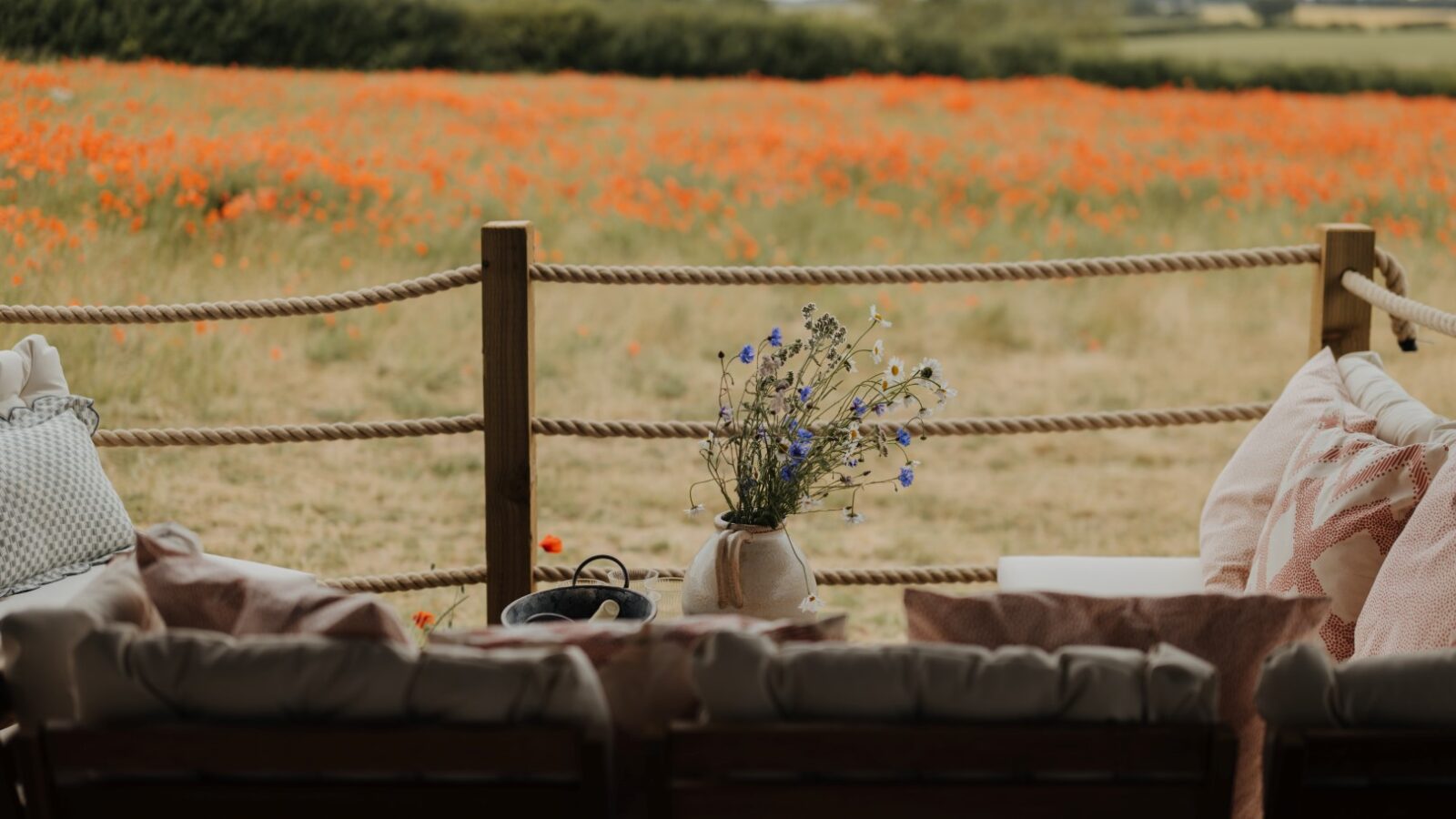 A cozy outdoor seating area with cushioned chairs is shown on a wooden deck at Westgate Farm. A vase with wildflowers sits on a small table. Beyond the deck, there is a field filled with vibrant orange flowers and dense green trees in the background.