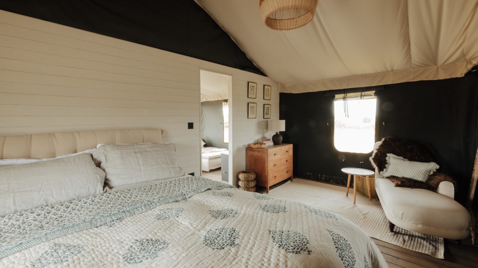 A cozy bedroom at Westgate Farm features a large bed adorned in white and patterned bedding, a wooden dresser with framed pictures above it, and a white armchair with a fluffy brown throw. A small window lets in natural light, and there's a doorway leading to another room.