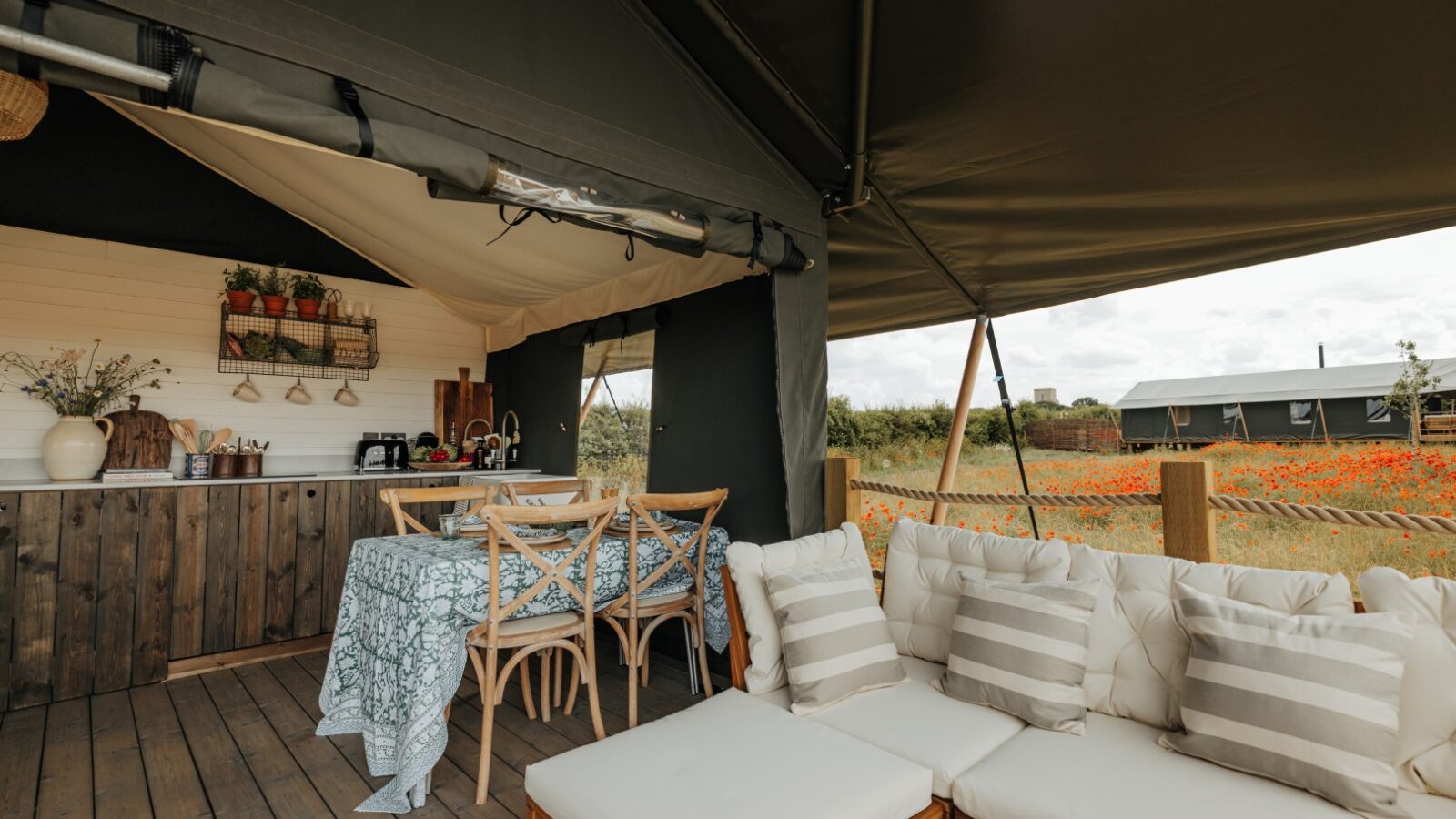 An outdoor dining and lounging area in a tent features a wooden table with four chairs, a kitchenette along the back wall, and a cushioned seating area with striped pillows. A second similar tent is visible in the background at Westgate Farm, surrounded by a field of orange flowers.