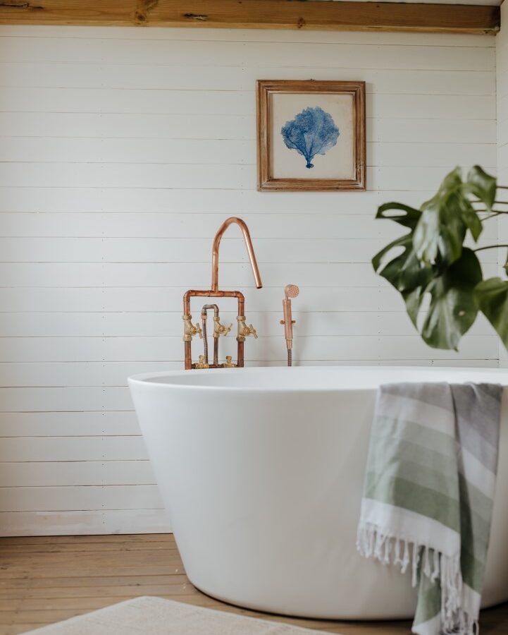 A modern bathroom at Westgate Farm features a white freestanding bathtub with copper fixtures. A green and white striped towel hangs over the tub's side. A framed blue coral artwork adorns the white wooden wall. A potted plant adds a touch of greenery, and a beige rug lies on the wooden floor.