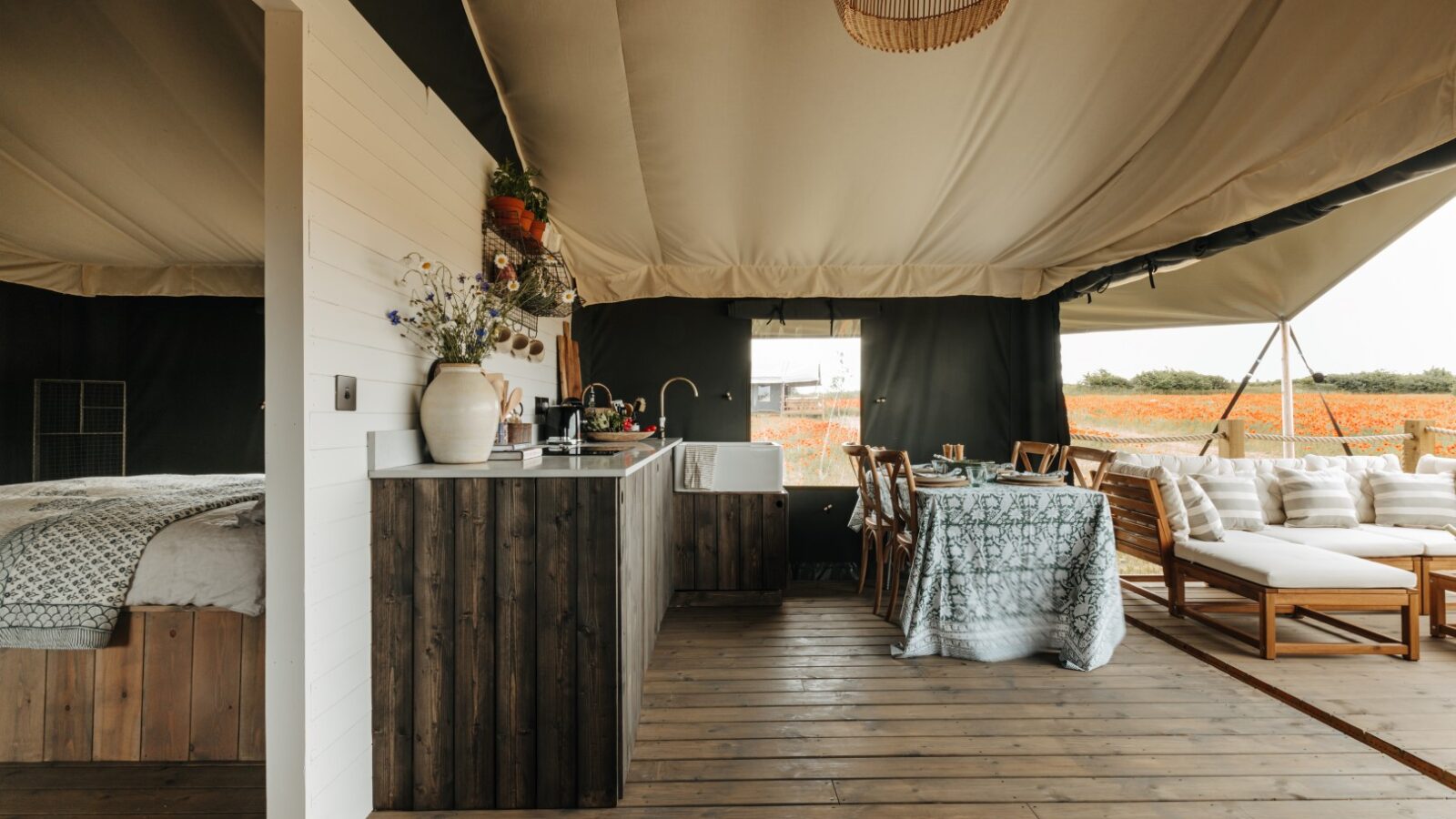 A modern, luxurious glamping tent interior at Westgate Farm features a wooden floor, a kitchenette with a dark wood countertop, a cozy dining area with a patterned tablecloth, and a lounge area with white cushioned seating. A bed is partially visible in a separate area.