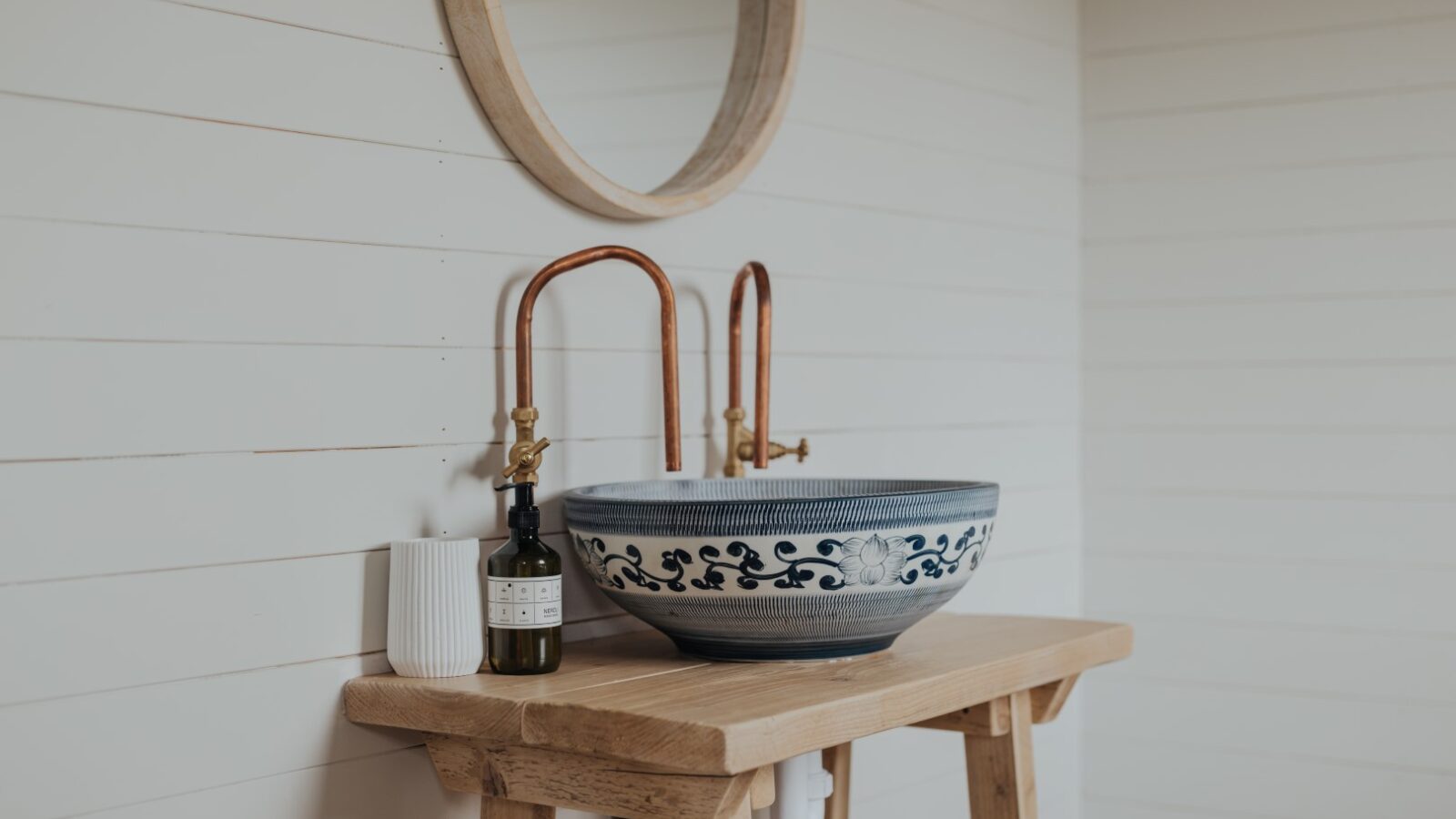 A wooden bathroom vanity from Westgate Farm features a decorative ceramic sink with blue floral patterns. The mirror above has a wooden frame, complemented by copper faucets. A bottle of soap and a white toothbrush holder are placed on the vanity, set against a wall with white horizontal panels.