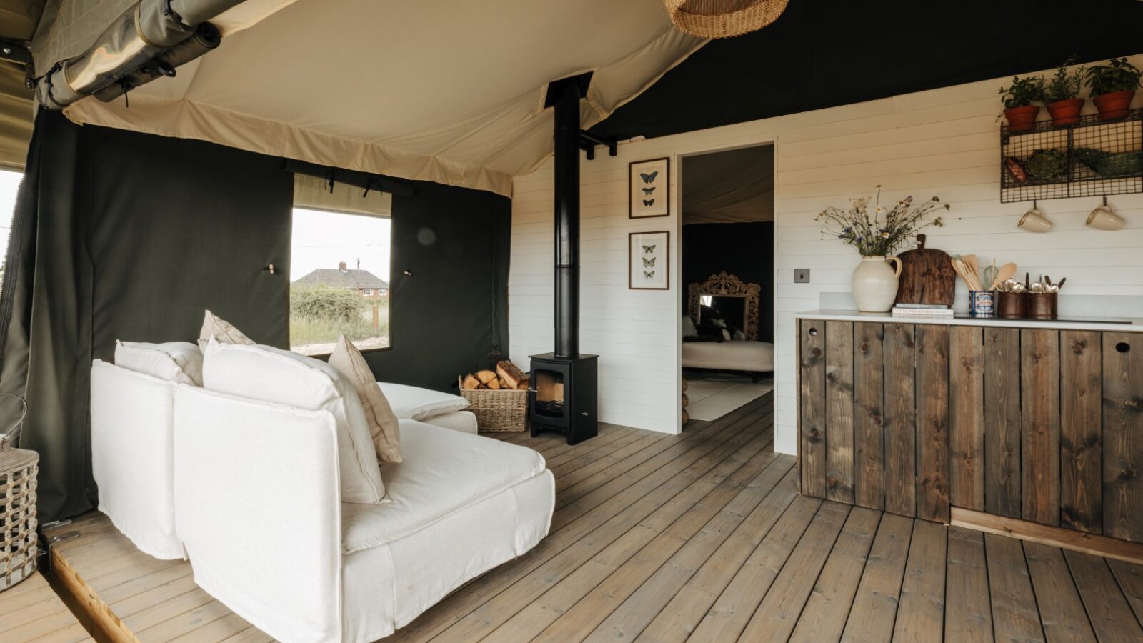 A cozy glamping tent interior at Westgate Farm features a white armchair, a wooden stove with a woodpile beside it, and a rustic wooden kitchen counter. The flooring is wooden, and there's a basket of firewood. The walls are adorned with butterfly prints and potted plants.