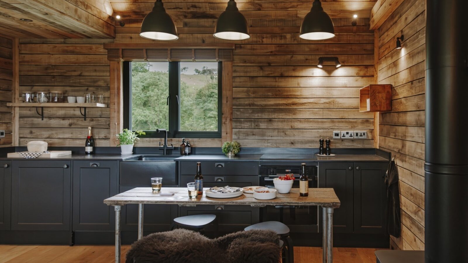 A cozy, rustic kitchen with wooden walls and ceilings reminiscent of classic wood cabins. It features modern, dark gray cabinetry, black pendant lights, and a wooden island with two stools. Dishes and bottles rest on the island, while a window offers a view of greenery outside.