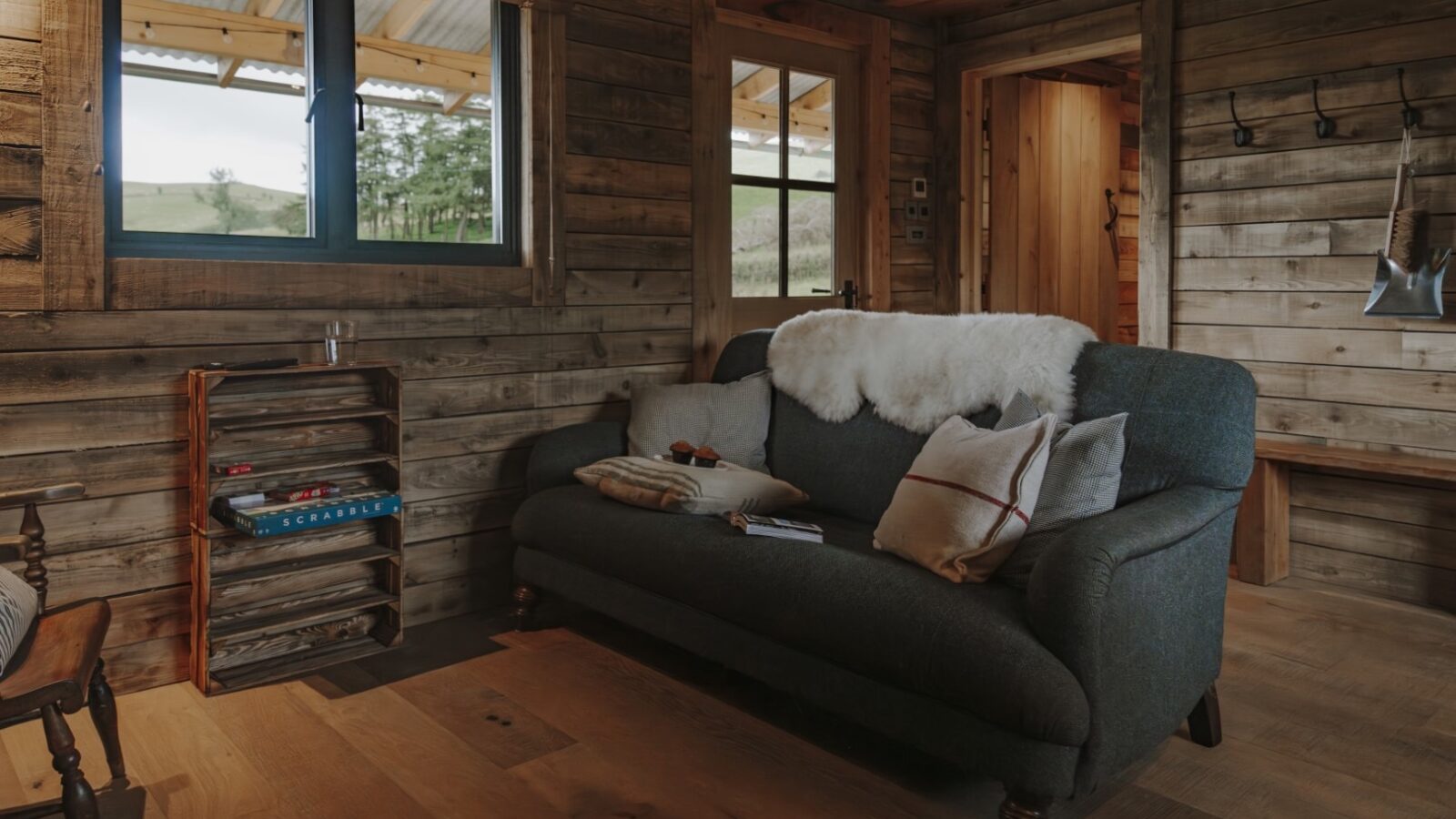 A cozy cabin living room featuring a dark green sofa with cushions and a white woolly throw, a wooden bookshelf with books and a lamp, and a window offering a scenic view. Wooden walls and floors create a rustic ambiance. Hooks and an axe are mounted on the wall by the door.