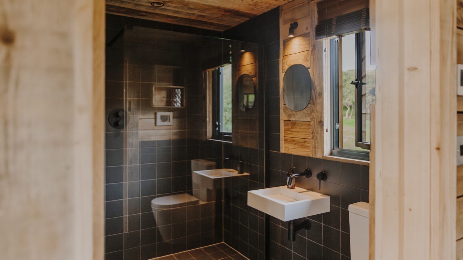 A modern bathroom in Woolly Wood Cabins features wooden walls and ceilings, a glass-enclosed shower, a white sink with a round mirror above it, and a toilet. Light streams in through a window, illuminating the tiled floor and dark wall tiles.