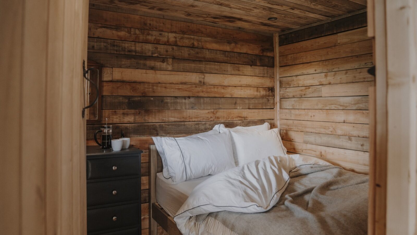 A cozy wooden bedroom with a rustic cabin feel. The bed is neatly made with white linens and a gray blanket. A small black nightstand beside the bed holds a woolly white mug and a glass jug. The walls and ceiling are crafted from light and dark wooden planks, enhancing the serene cabin ambiance.