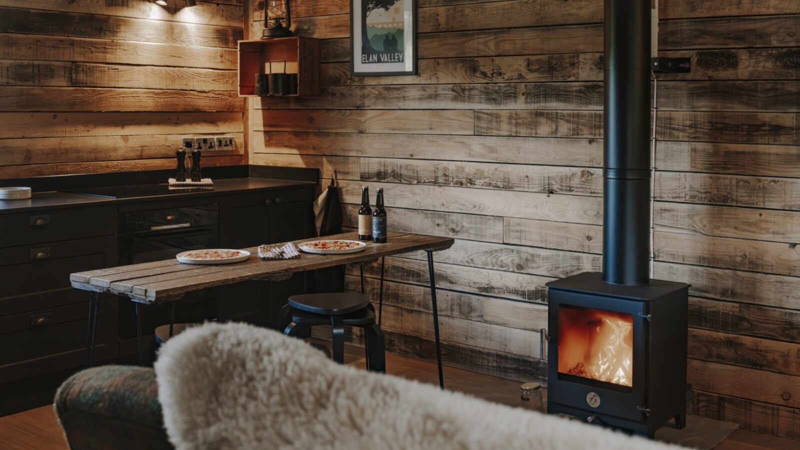 A cozy cabin interior at Woolly Wood Cabins features wooden walls, a small black wood-burning stove with a glass door, a rustic wooden table set with plates of food, and dark cabinetry. Warm lighting adds a welcoming ambiance, and a furry blanket drapes over a chair in the foreground.