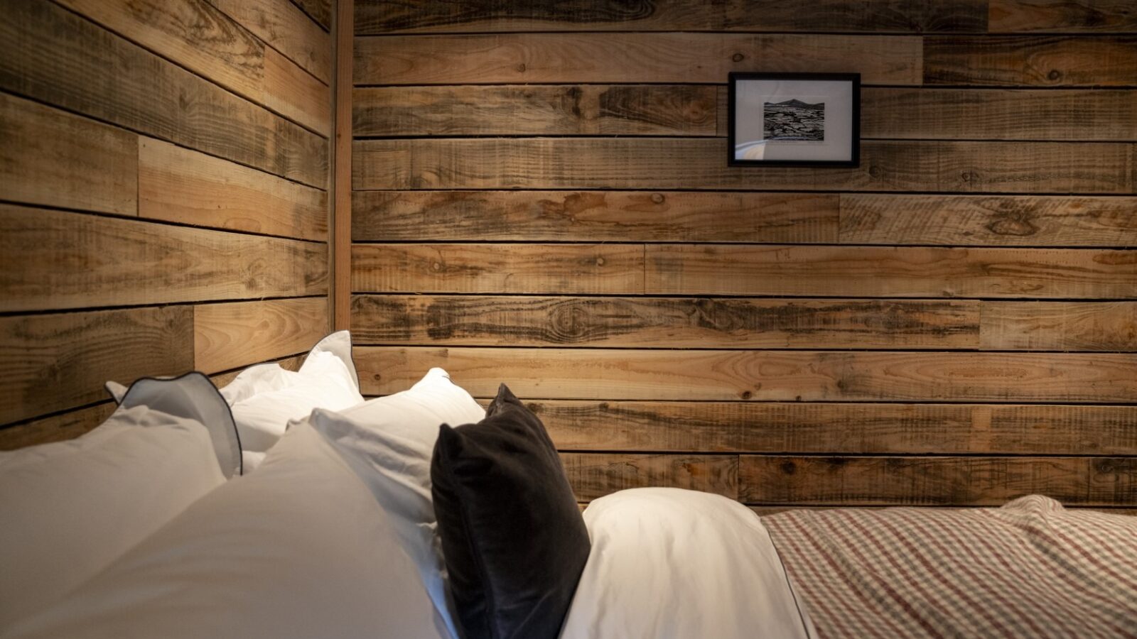 A cozy bedroom with wooden paneled walls, reminiscent of rustic cabins. The bed, adorned with white pillows and a gray pillow, boasts a multi-patterned blanket that adds charm. A small framed black-and-white photo hangs above the bed, completing the inviting woolly atmosphere.