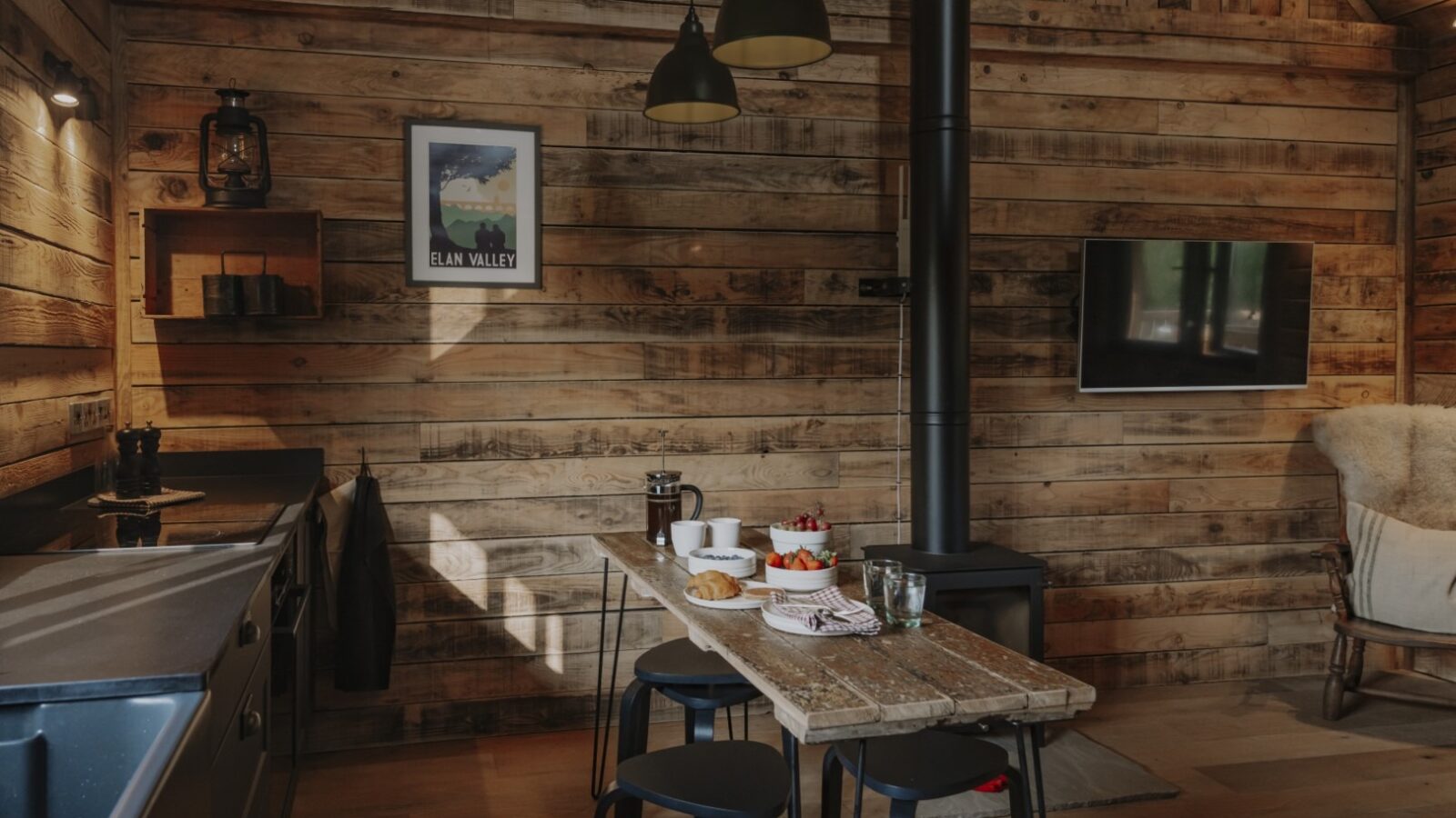 The cozy kitchen at Woolly Wood Cabins boasts rustic wooden walls, a modern stove, and a central dining table with black stools. Set for breakfast with pastries and fruit, the scene is complete with a framed picture above a shelf and a flat-screen TV mounted nearby.