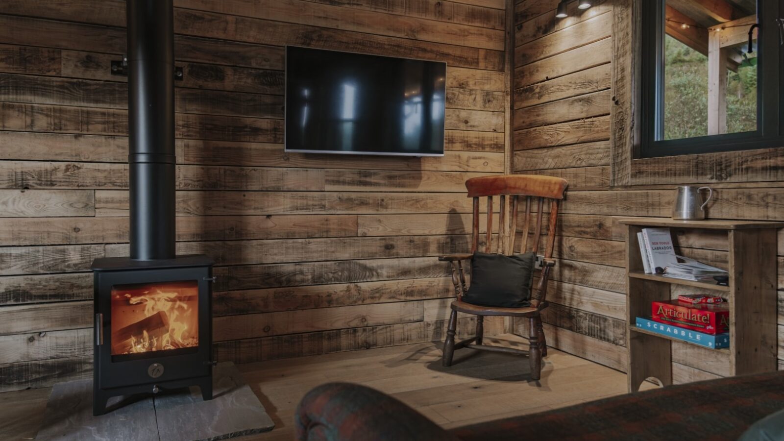 The cozy interior of Woolly Wood Cabins features wooden walls, a lit wood-burning stove, a flat-screen TV mounted on the wall, a rocking chair, and a small side table holding books and a mug. The room exudes a warm, inviting ambiance with soft lighting and rustic wooden elements.