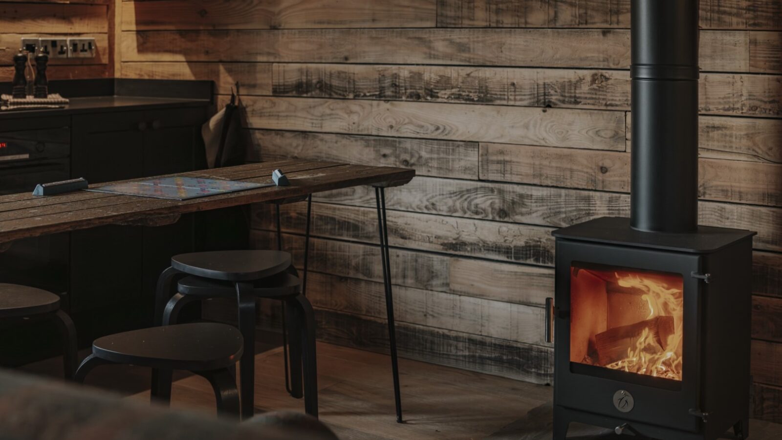         A cozy indoor setting at Woolly Wood Cabins features a wood-burning stove with a visible flame, exuding warmth. The space has wooden walls and floors, a rustic table with black stools, and a dark-toned kitchen counter in the corner. The ambiance is warm and welcoming.