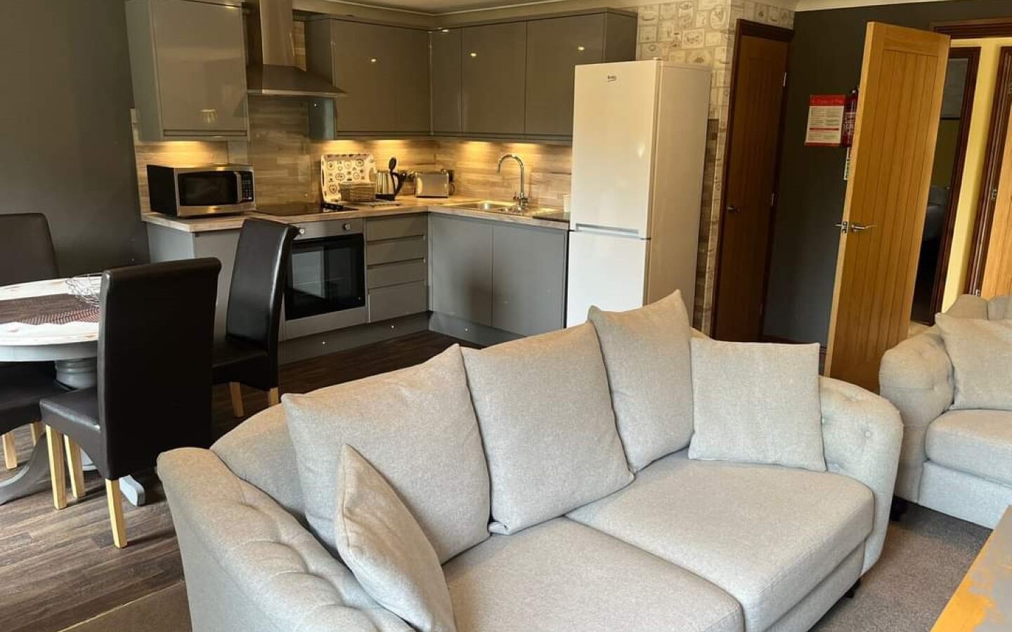 A modern kitchen and living area with gray cabinets, stainless steel appliances, and a white refrigerator. The space includes a light gray sofa, a dining table with black chairs, and wooden flooring. Visible through the partially open door is a glimpse of Country Park's serene greenery.