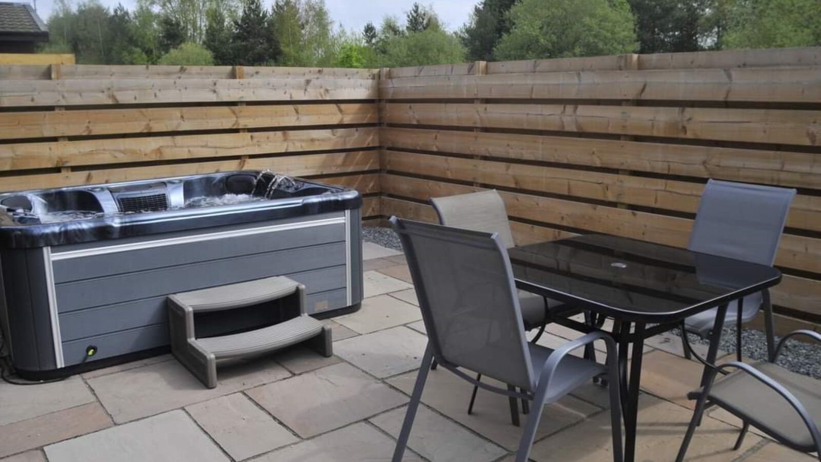 A backyard patio area features a hot tub with a small step, surrounded by a tall wooden fence. Next to the hot tub is a glass-top table with four chairs. The patio surface is made of stone tiles, and greenery reminiscent of Country Park is visible beyond the fence.