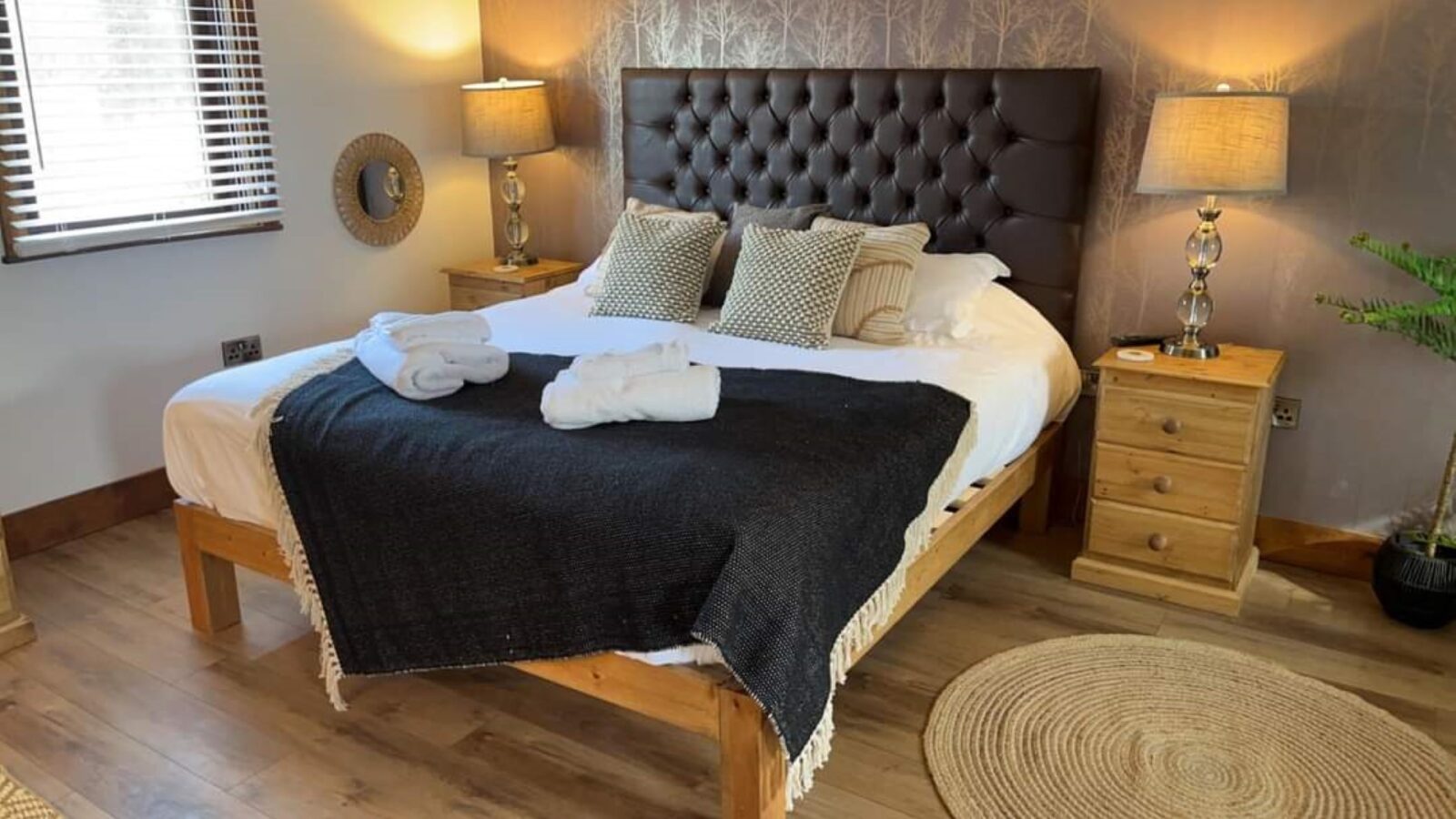 A cozy bedroom with a wooden floor and a large bed. The bed has a dark headboard, white bedding, and a black throw blanket. Two rolled towels rest on the bed. Wooden bedside tables with lamps flank either side, while a window with wooden blinds lets in natural light, offering views of Country Park.