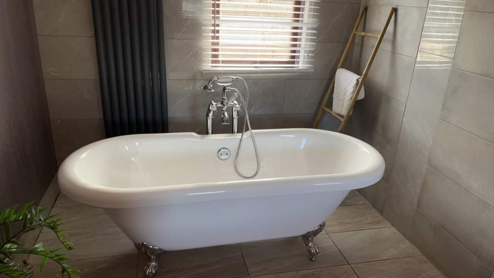 A modern bathroom in Country Park features a white clawfoot bathtub with a silver faucet and showerhead. Behind it is a window with blinds, and to the right, a wooden ladder rack holding a white towel. A green plant rests on the left side. The decor is in neutral tones.