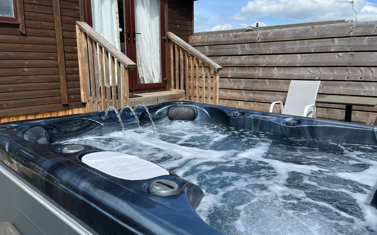 A bubbling hot tub with jets is placed outdoors on a wooden deck next to a wooden cabin in Country Park. A white lounge chair is positioned beside the tub, and a tall wooden fence surrounds the area. The sky is clear with a few clouds.