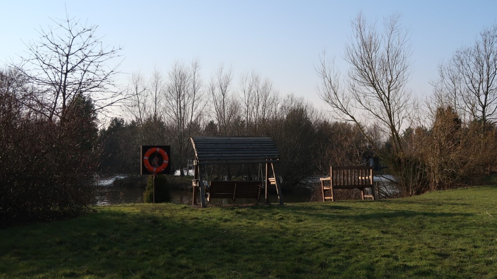 A picturesque lakeside scene at Country Park features wooden benches and a covered wooden swing facing the water. Leafless trees and bushes surround the calm lake, while a life preserver is mounted on a post beside the swing. Grass covers the foreground, illuminated by afternoon light.