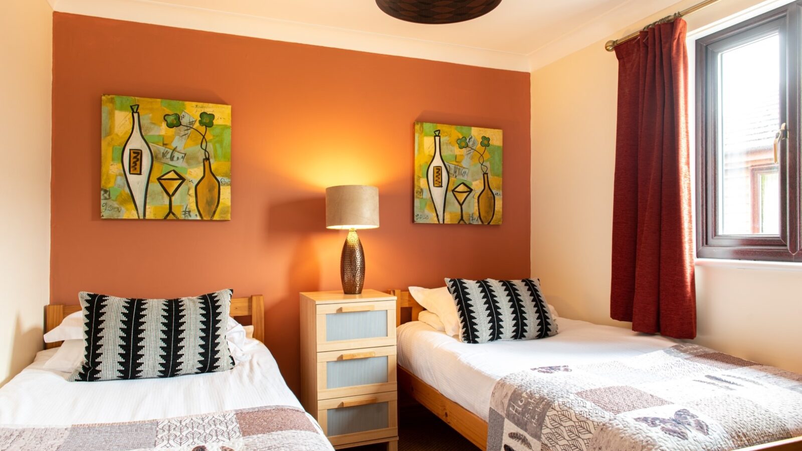 A cozy room with two single beds, each adorned with patterned pillows and blankets. A wooden nightstand with a modern lamp is placed between the beds. The walls are painted a warm orange and cream, decorated with two abstract paintings inspired by Yellowtop Country Park. A window with red curtains is on the right.