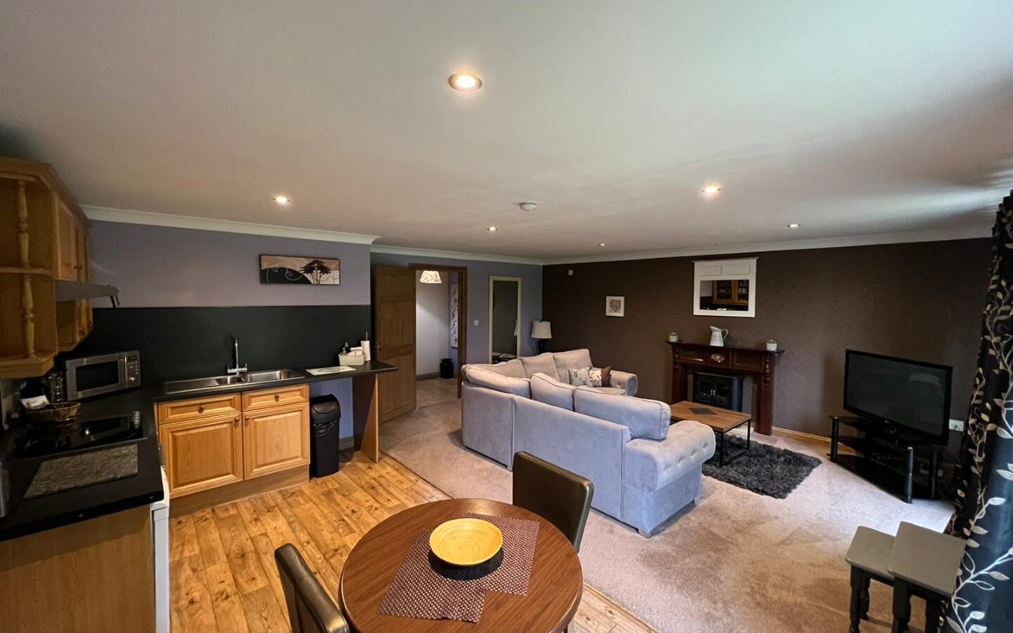 A spacious open-plan living area with a kitchen, dining table, and seating area. The kitchen features wooden cabinets and a Yellowtop countertop. The living area has a gray sectional sofa, TV, and a fireplace. Large windows provide ample natural light, highlighting the wooden floors with views of Country Park.