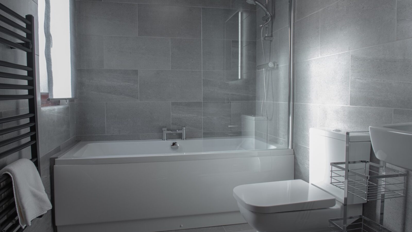 A modern bathroom with gray tiles, featuring a bathtub with a glass shower screen, a white toilet, a white sink, and a metal storage rack. A black heated towel rail with a white towel hangs on the left wall. Natural light from the window highlights views of nearby Country Park.