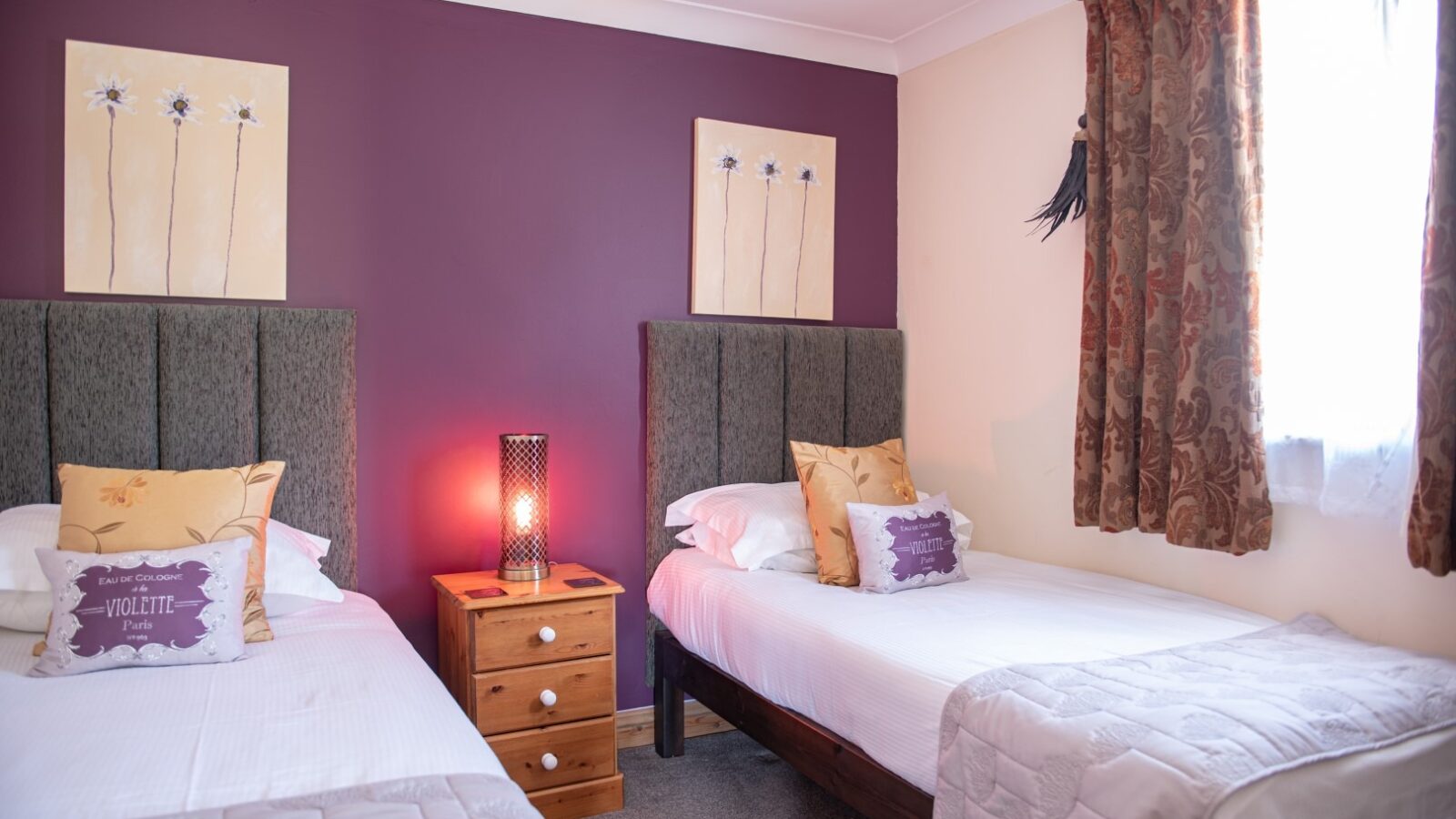 A cozy bedroom with two single beds adorned with white bedding and accent pillows. The room features a nightstand with a lit lamp between the beds, purple accent walls, floral paintings above each bed, and a window with patterned curtains letting in natural light, evoking the charm of Country Park.