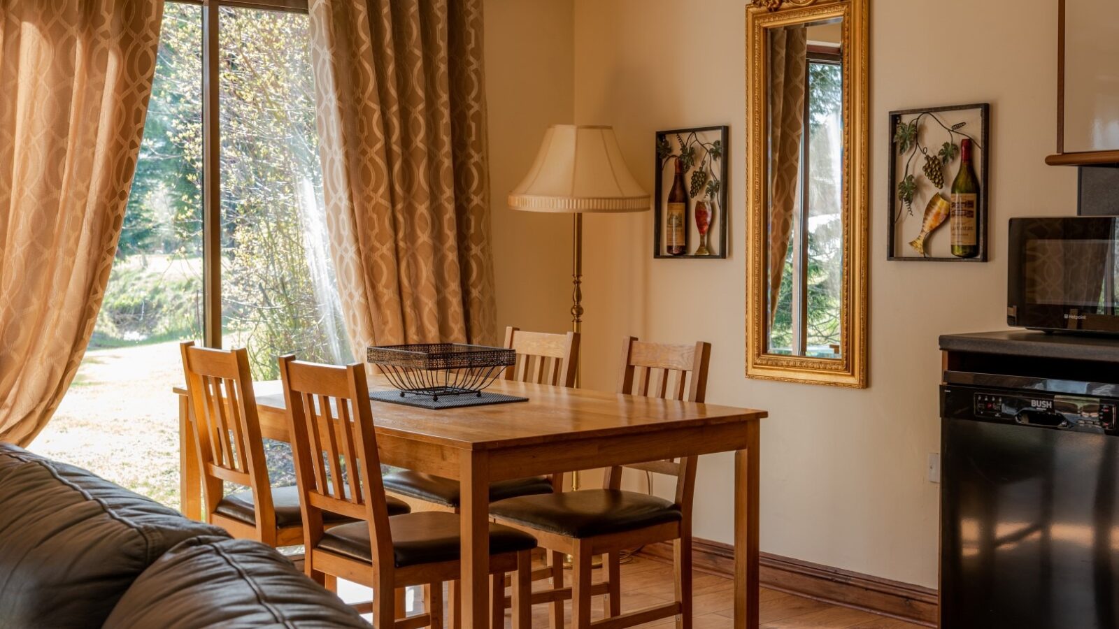 A cozy dining area with a wooden table and four chairs, positioned near a window with beige curtains. The wall features a tall decorative mirror and two framed pictures of wine bottles, reminiscent of Country Park charm. A lamp stands beside the table, and a black appliance is partially visible.