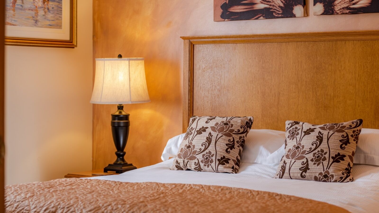 A cozy bedroom with a neatly made bed featuring a light brown bedspread and two decorative pillows with floral patterns. A bedside table with an elegant lamp is next to the bed. Above the bed, there's wall art and a framed picture of Yellowtop Country Park on the adjacent wall.