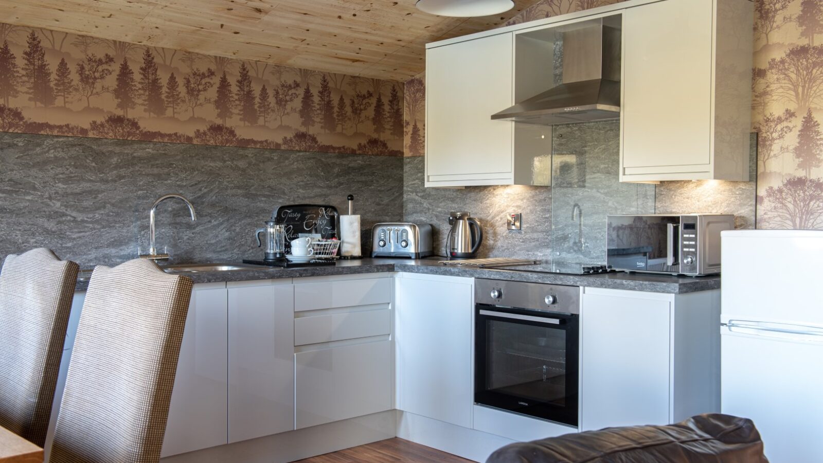 Modern kitchen with wood-paneled ceiling and tree-themed wallpaper, reminiscent of Yellowtop Country Park. It features white cabinets, a stainless steel range hood, an oven, a microwave, a toaster, a coffee maker, and a kettle. Two dining chairs and part of a brown sofa are visible in the foreground.