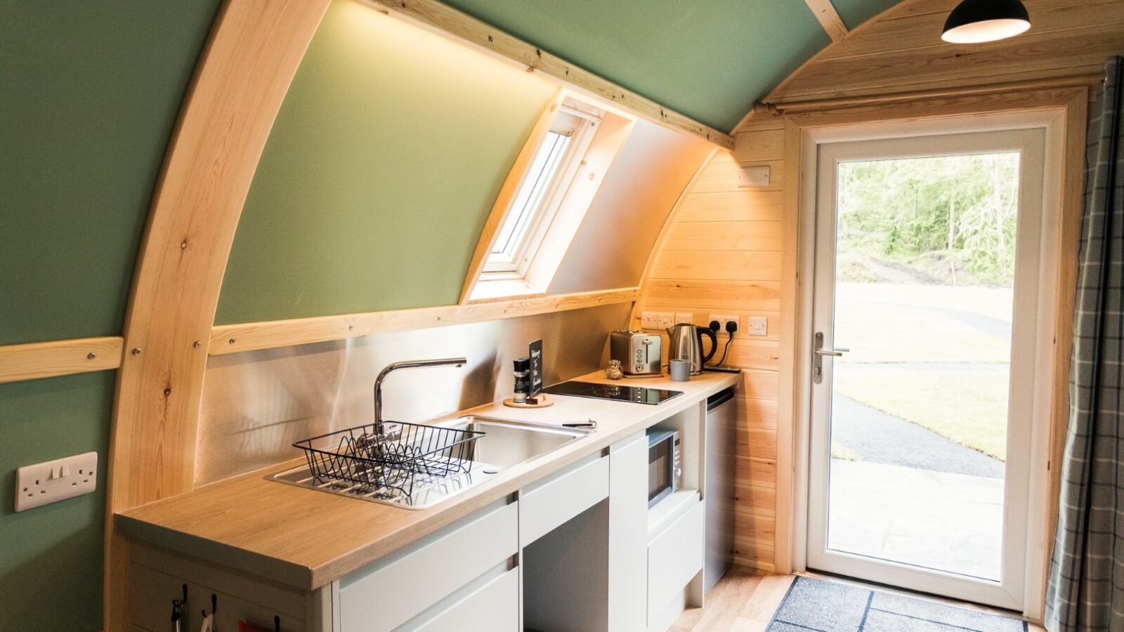 A small, modern kitchen with wooden finishes inside a curved, cabin-like structure at Forcett Grange. It features a sink, countertop, stove, kettle, and a coffee machine. There's a window above the sink and a door with a glass panel leading outside. Natural light illuminates the space.