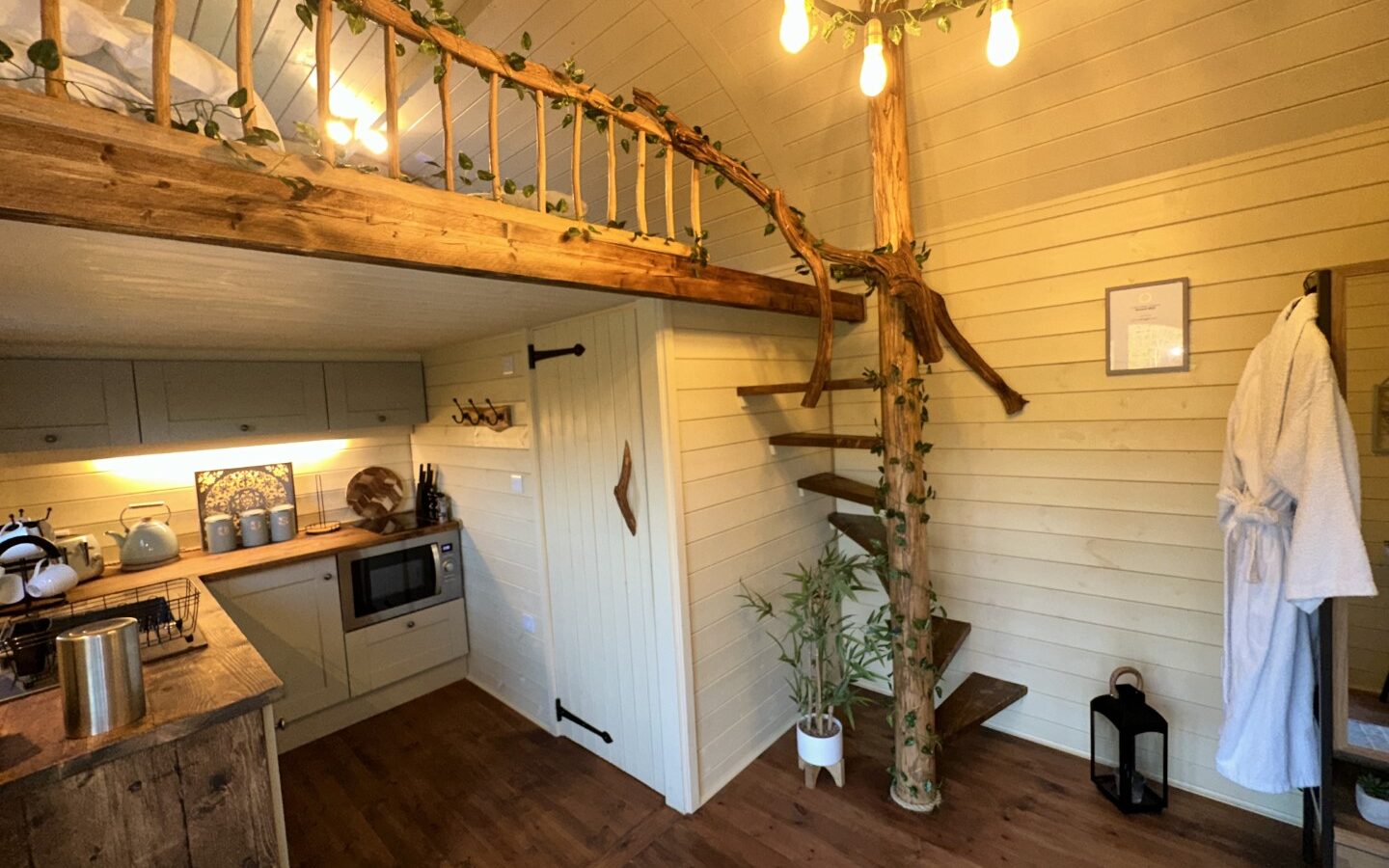 A cozy, rustic tiny home interior at Freedom Fields Glamping features a wooden staircase leading to a loft. The kitchen area offers light-colored cabinets, a microwave, and various utensils. A wooden beam entwined with greenery supports the loft, and warm lighting adds to the inviting ambiance.