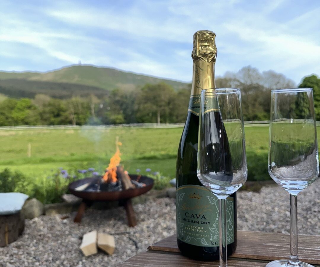 A bottle of Cava and two empty champagne glasses sit on a wooden table in an outdoor glamping setting. In the background, Freedom Fields boasts a fire pit with a small fire, green fields, trees, and a hilly landscape under a partly cloudy sky.