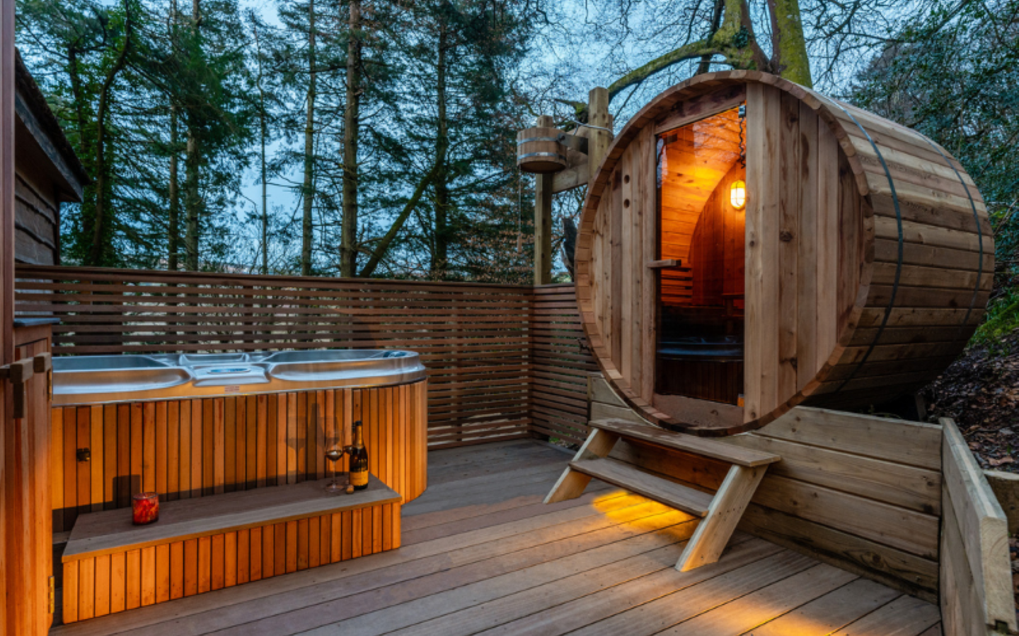 The image shows a cozy outdoor spa area at Ravendere Treehouse with a wooden barrel sauna and an adjacent hot tub. The sauna has a warm interior light, and the wooden deck is illuminated with soft ambient lighting, set against a backdrop of trees, creating a serene natural setting.