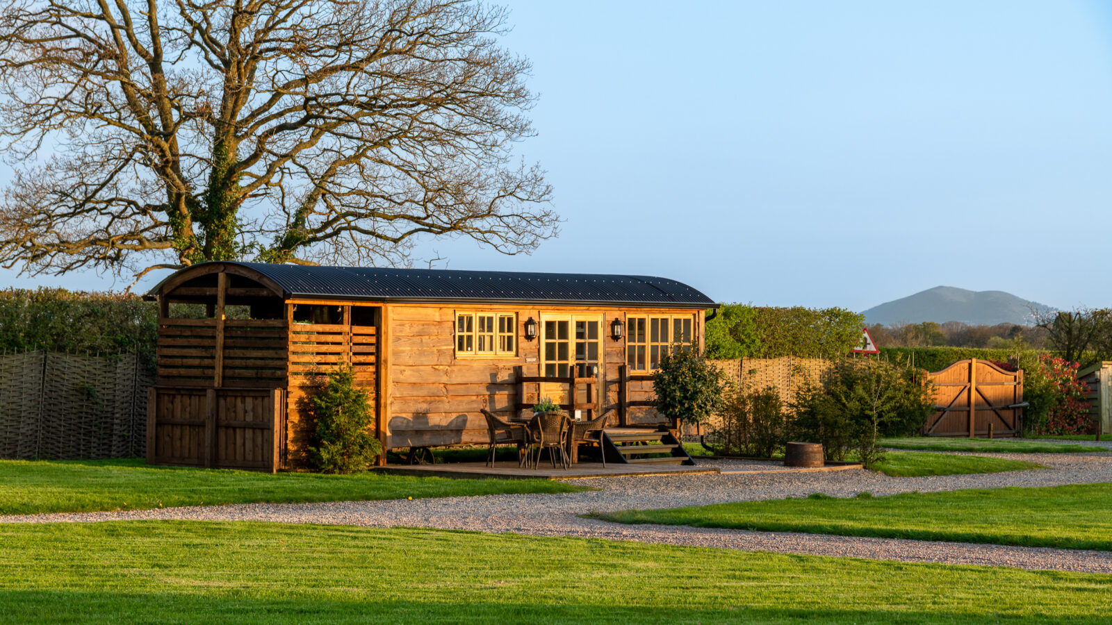 A charming wooden cabin with a small deck sits on a well-manicured lawn at Ockeridge Rural Retreats. The cabin features a glass door and windows, a side shed, and outdoor seating. A large leafless tree stands behind it, with a mountain under the clear, blue sky in the background.