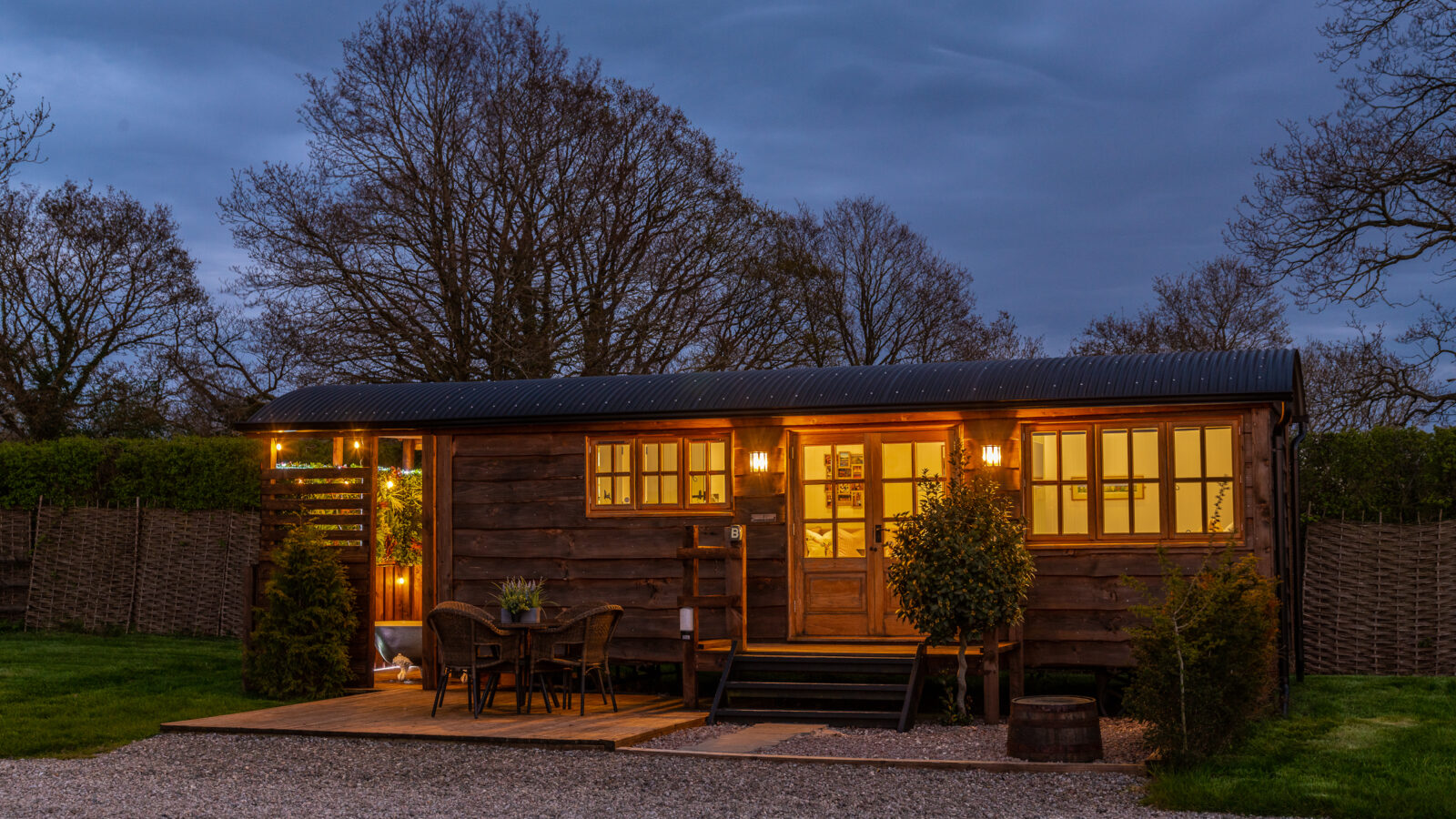 The image shows a cozy, rustic cabin at dusk. Warm yellow lights illuminate the wooden exterior and porch. A table with chairs, plants, and a small tree decorate the porch. This serene rural retreat is set against a backdrop of leafless trees, with a gravel pathway in the foreground.