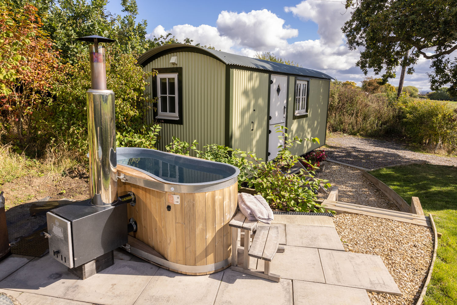 A luxury shepherds hut with white trim stands on a gravel path. In the foreground, there's an outdoor hot tub with a wood-burning stove heater beside it. This luxury shepherd's hut is surrounded by trees and bushes, and the sky is partly cloudy.