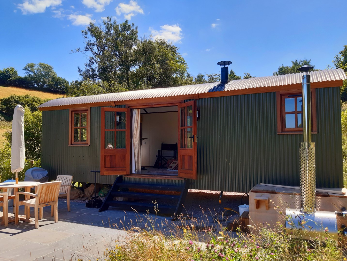 A green, luxury shepherds hut with wooden doors and windows sits on a sunny day. The doors are open, revealing a cosy interior. There's a small outdoor seating area with a table, chairs, hot tub and a barbecue grill. Trees and a grassy field surround the shepherds hut.