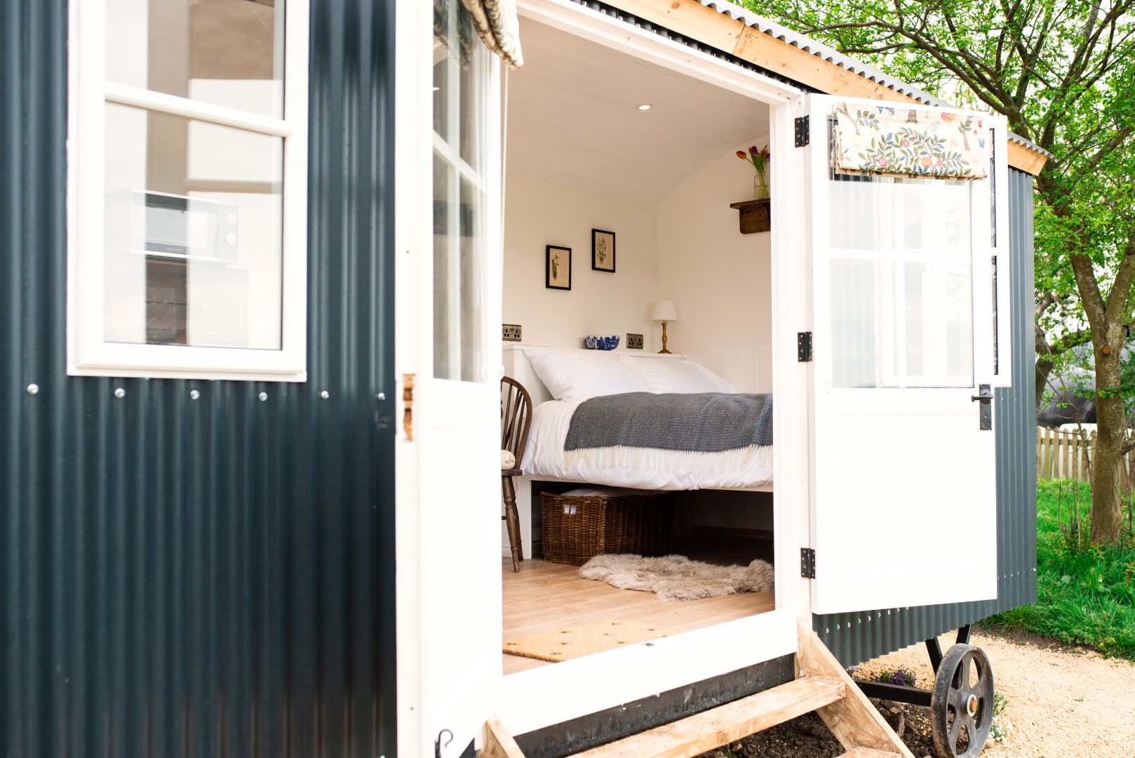 A luxury shepherd's hut with black and white exterior walls and a covered entrance. The wooden steps lead to an interior featuring a cozy bed with white and gray linens, a wooden chair, a nightstand with a lamp, and framed pictures on the walls. 