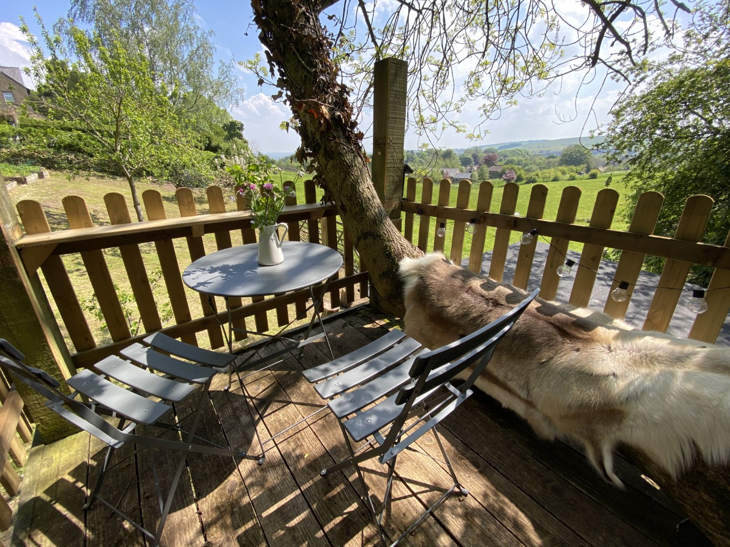 A cozy outdoor seating area with a grey metal table and two matching chairs on a wooden deck, perfect for enjoying a gin on a summers evening. Cloesby is your luxury shepherds hut accommodation. The deck is surrounded by a wooden fence, and a tree trunk leans against it. A vase with flowers sits on the table, offering views of greenery and hills.